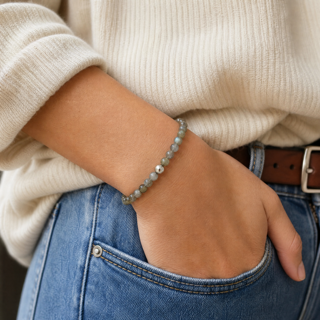 A close-up of a person wearing a beaded bracelet on their wrist, paired with a cream ribbed sweater, blue jeans, and a brown leather belt.