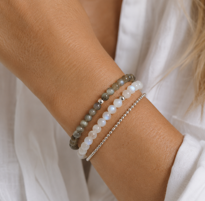 A close-up of a female model's wrist wearing three beaded bracelets: a grey beaded labradorite gemstone bracelet, a white rainbow moonstone beaded bracelet, and a thin silver chain bracelet, styled with a white top.